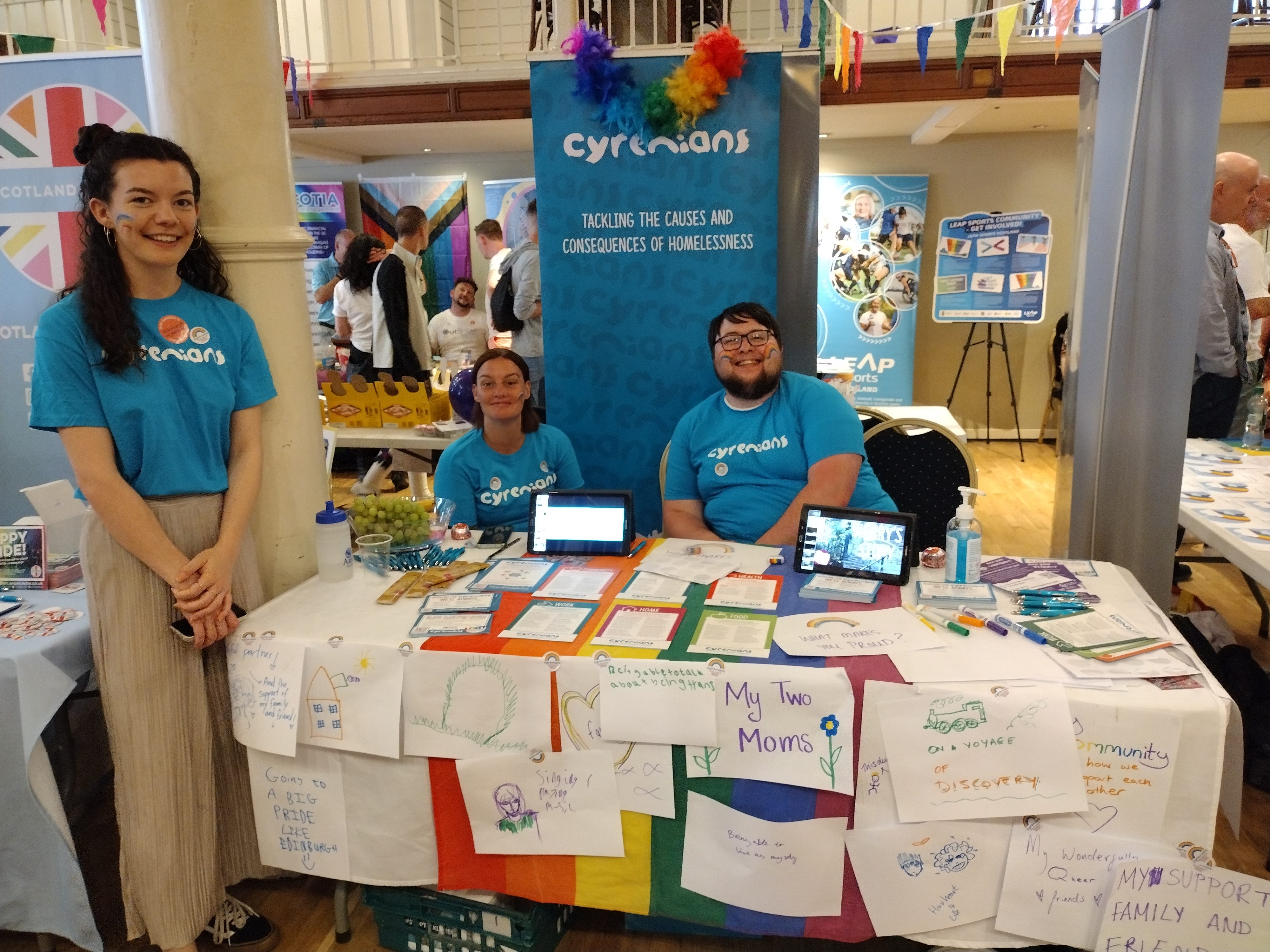 A rainbow patterned stall at Pride 2022 manned by 3 people in Cyrenians tshirts